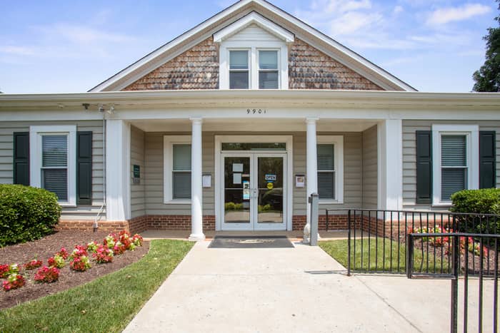 Front exterior of a single-story community clubhouse numbered 9901 with cedar-shake gable, white columns flanking glass double doors, a flowering flower bed, and a paved walkway leading to the entrance.