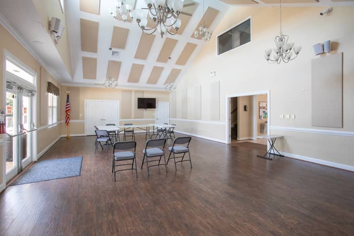 Interior community room with a high coffered vaulted ceiling, two chandeliers, hardwood-look flooring, folding chairs and tables, a mounted TV, an American flag, and glass doors letting in daylight.