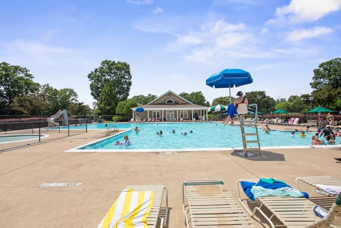 Large outdoor swimming pool with people swimming, a lifeguard chair under a blue umbrella, lounge chairs and pool umbrellas around the deck, a water slide, and a pool house building beneath a blue sky.