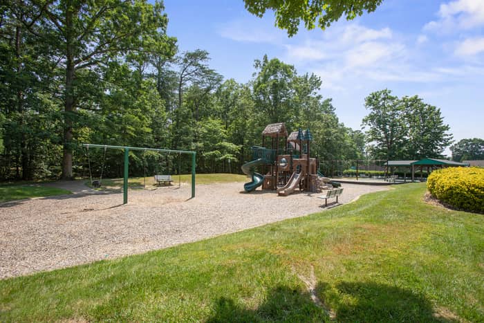 Tree-lined community playground with a wooden play structure featuring a green spiral slide, a metal swing set, benches on a wood-chip surface, and a covered shelter in the background beside a grassy lawn.