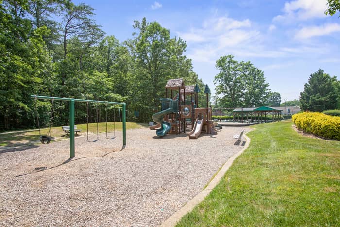 Community playground with a wooden climbing structure and green spiral slide, a metal swing set with several swings, benches, a wood-chip ground surface, and a covered pavilion behind the trees.