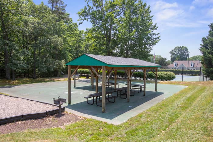 Open-sided wooden pavilion with a green-and-shingle roof shading several picnic tables on a concrete pad, a charcoal grill at the edge, set among trees with tennis courts and houses in the background.