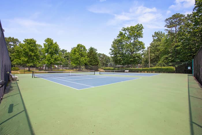 Blue-and-green hard-surface tennis courts with nets in place, enclosed by chain-link fencing, bordered by mature green trees under a blue sky.