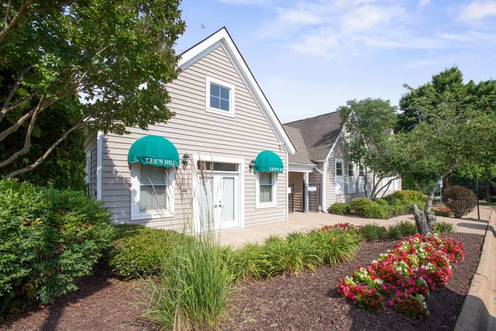 Two-story gray clubhouse building with green Lee's Hill awnings over the doors and windows, surrounded by manicured landscaping, ornamental grasses, and red flowering shrubs along a paved walkway.