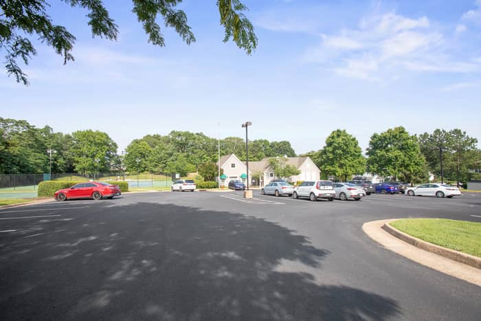 Paved community parking lot with several parked cars in front of a clubhouse building, with tennis courts visible to the left and mature trees lining the property under a partly cloudy sky.