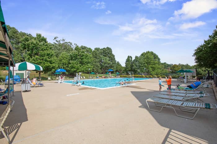 Large outdoor community swimming pool with a sunny concrete deck, green-and-white striped umbrellas, lounge chairs, and a few swimmers, framed by mature trees under a blue sky