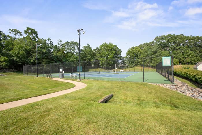 Fenced multi-court tennis complex with green and blue playing surfaces, court lighting, and a bench, set beside a curving paved path and grass lawn ringed by trees
