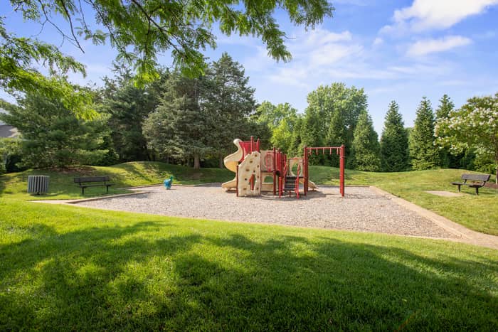 Children's playground with a red and tan climbing structure, slide, and swing set on a wood-chip pad, flanked by benches and a trash bin, surrounded by grass and evergreen trees