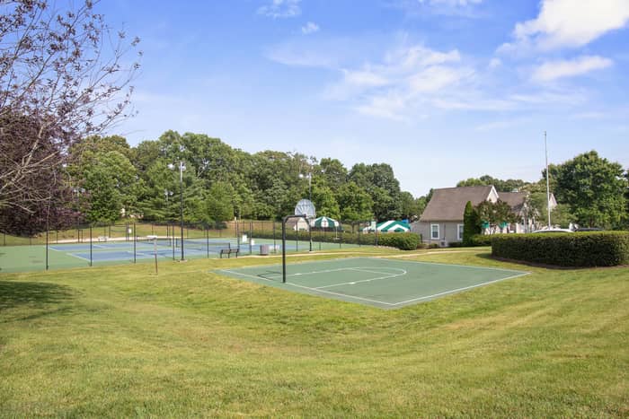 Green half-court basketball court with hoop in the foreground, tennis courts and a clubhouse-style building visible behind across a grass lawn under a partly cloudy sky