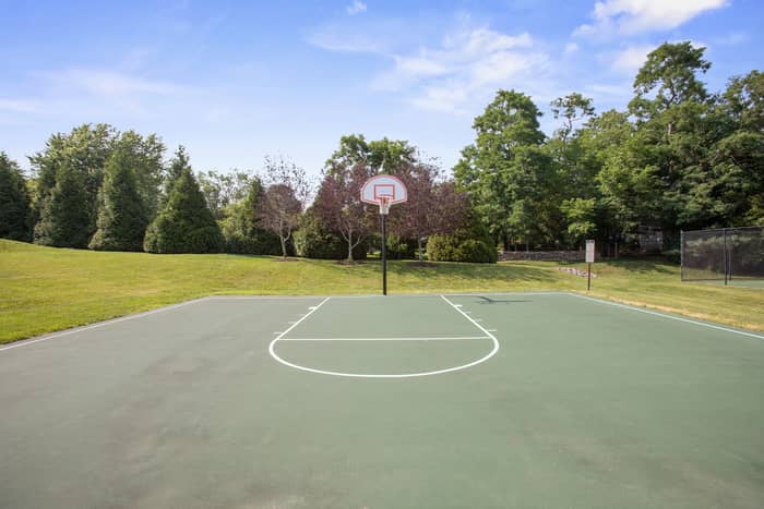 Green outdoor basketball court with freshly painted white key and three-point lines, a single hoop, and a second pole, backed by a manicured lawn, evergreens, and a stone wall