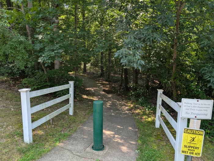 Paved walking trail entrance leading into shaded woods, with white split-rail fencing on both sides, a green bollard, and posted homeowner-use and slippery-when-wet caution signs