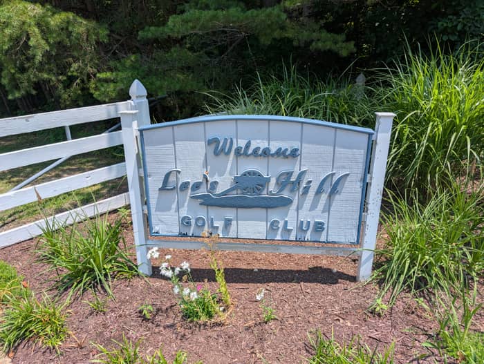 Weathered carved sign reading Welcome Lee's Hill Golf Club mounted on white posts, set in a mulched bed with wildflowers beside a white fence and tall grasses
