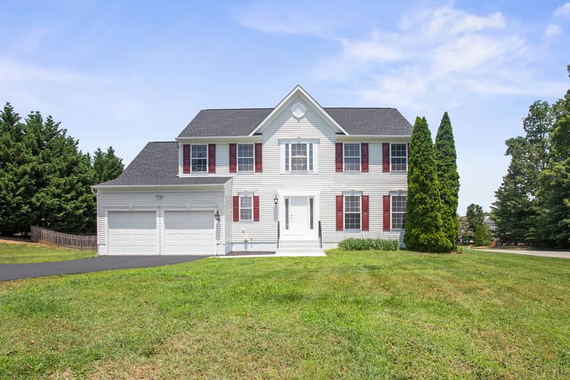 Front view of a two-story white vinyl Colonial home with red shutters, attached two-car garage, covered entry, and sloping green front lawn under a blue sky