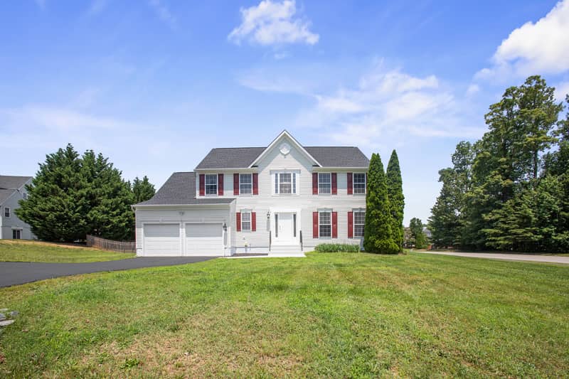 Angled front-left view of a two-story white Colonial home with red shutters, two-car garage, and driveway, set on a sloping lawn with neighboring homes nearby