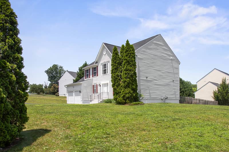 Side view of the white Colonial home showing the garage wing and tall gable end, framed by mature evergreen trees and an expansive green yard