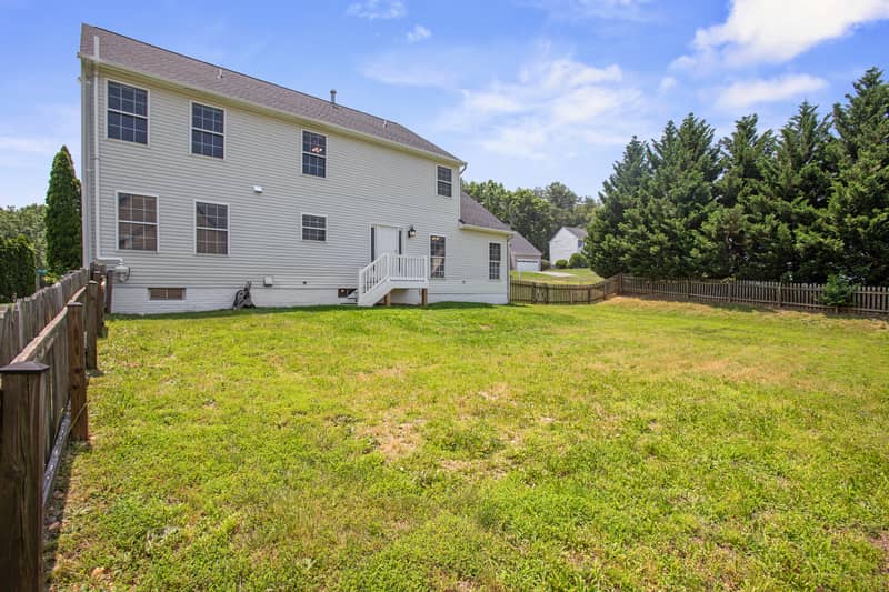 Rear view of the white Colonial home with a raised wood deck and stairs, surrounded by a large fenced grassy backyard and evergreen trees