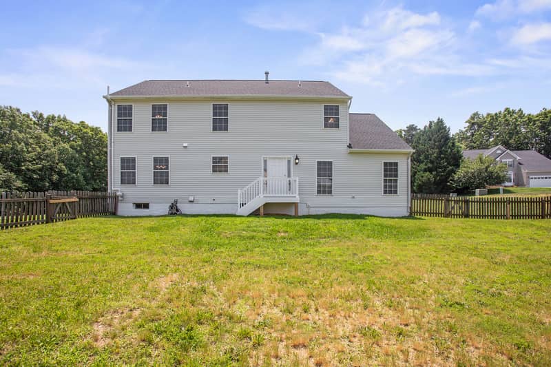 Symmetrical rear view of the white Colonial home with a raised deck and stairs, set behind a large fully fenced grassy backyard under a blue sky
