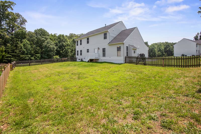 Wide view of the large fully fenced grassy backyard with the rear and side of the home at left and a tree line along the perimeter