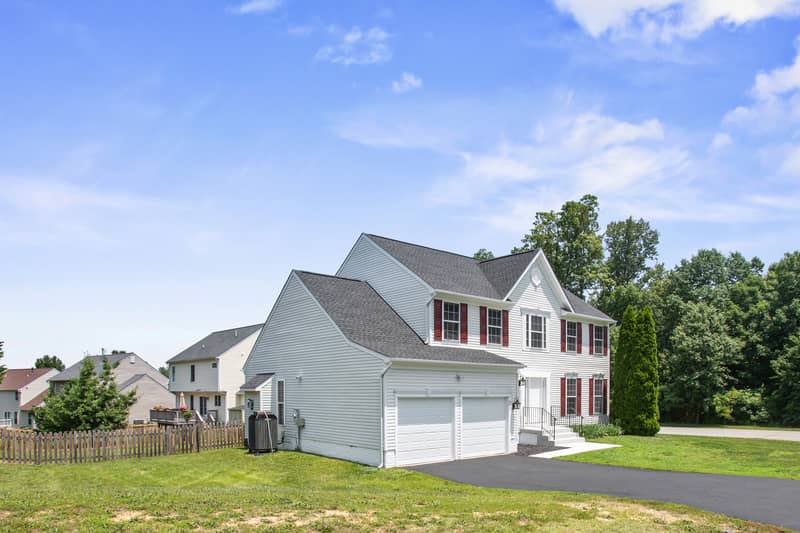 Wide angled view of the white Colonial home showing the garage-side elevation and front facade, with driveway, lawn, and neighboring homes under a blue sky