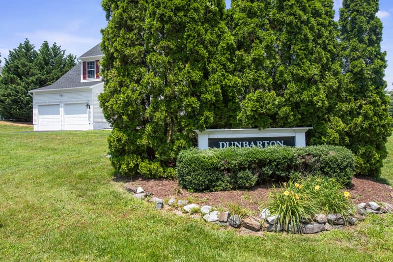 Landscaped Dunbarton neighborhood entrance sign surrounded by evergreens and daylilies, with the home visible in the background