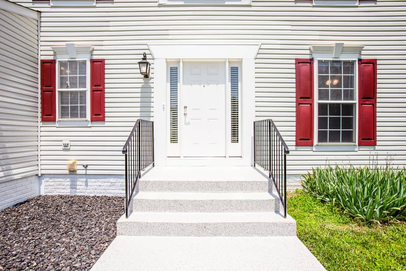 Close-up of the home's front entry showing a white six-panel door, carriage lamp, finished concrete steps with metal railings, and flanking windows with red shutters
