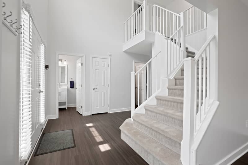 Entry foyer with sunlight through sidelight windows, dark wood-look flooring, a carpeted staircase with white balusters, and a glimpse of a powder room