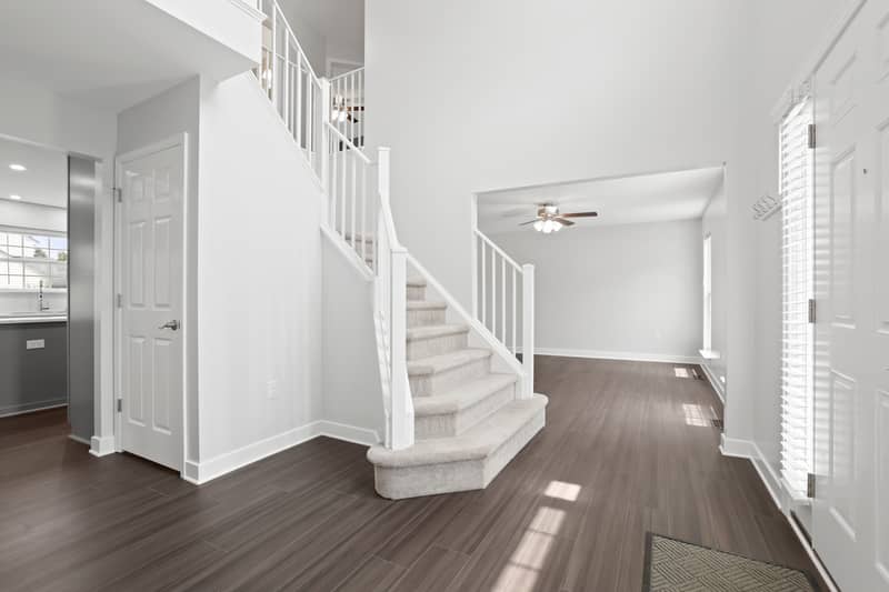 Foyer with carpeted staircase, dark LVP floors, an adjoining room with a ceiling fan, and a view into the kitchen with stainless appliances