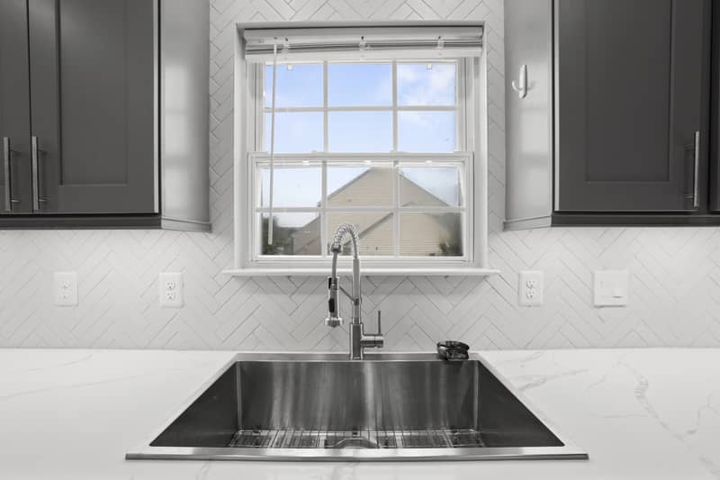 Kitchen with gray shaker cabinets, white quartz counters, stainless sink under a window, stainless range and dishwasher, herringbone tile backsplash