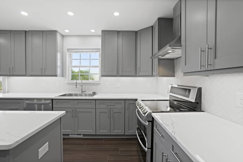 Open-concept kitchen with quartz island and gray cabinets flowing into a family room with a fireplace, large windows, and dark wood-look floors