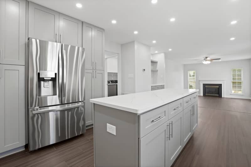 Open kitchen with floor-to-ceiling gray cabinetry, stainless steel French-door refrigerator, a long white quartz island, and recessed lighting, opening to a family room with a fireplace