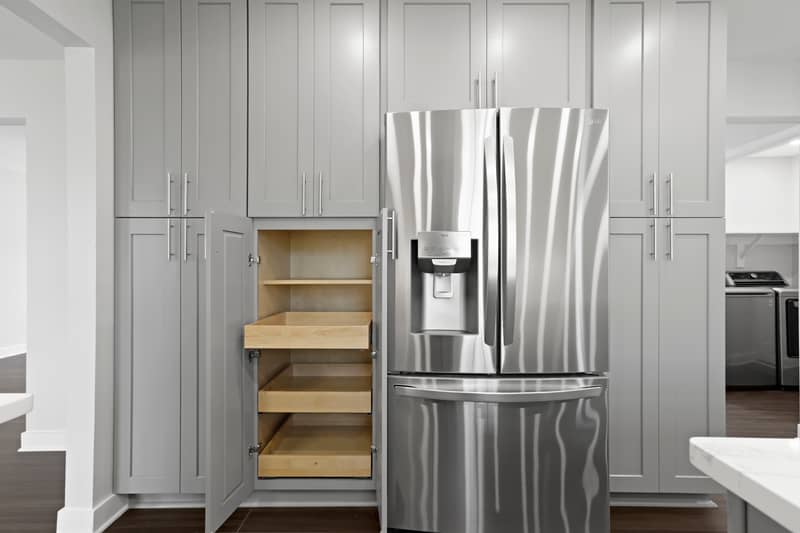 Detail of gray floor-to-ceiling kitchen cabinetry beside a stainless steel French-door refrigerator, with one cabinet open to reveal wood pull-out pantry shelves; laundry room visible at right