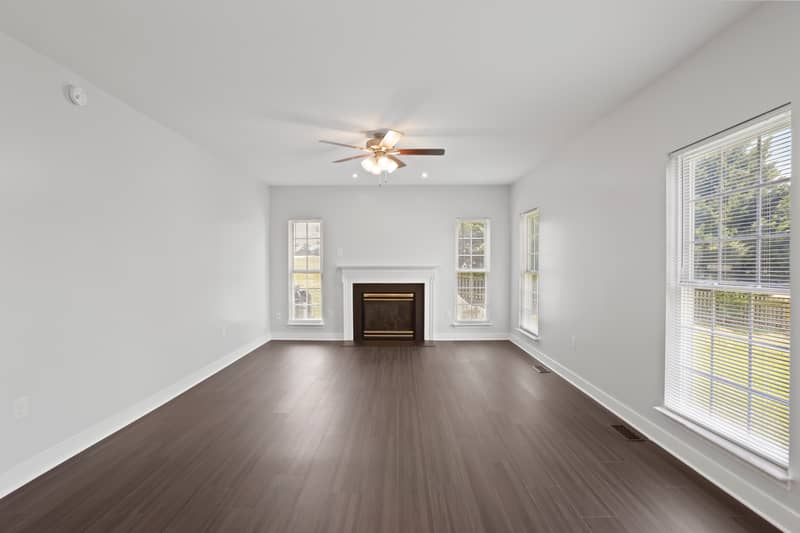 Empty family room with dark LVP flooring, a white-mantel gas fireplace flanked by windows, a ceiling fan, and a large window overlooking the fenced backyard