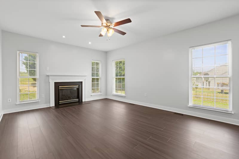 Empty family room with dark LVP flooring, white-mantel gas fireplace, ceiling fan, and several windows letting in natural light