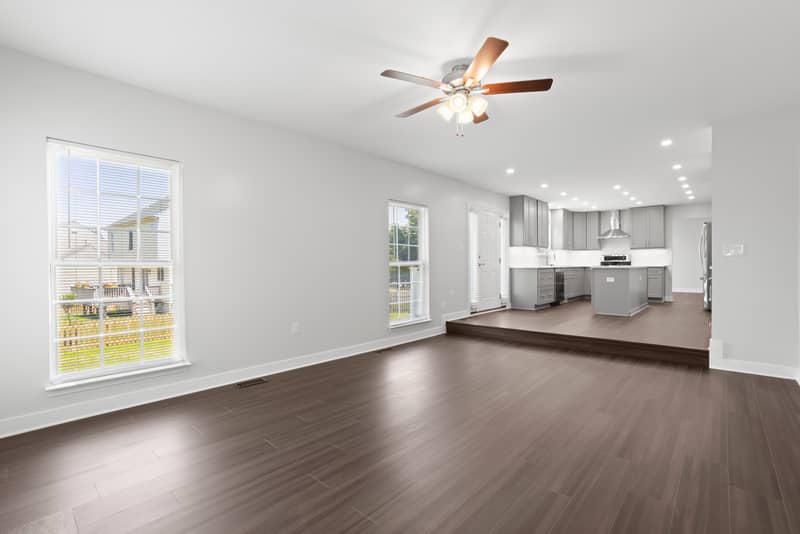 Spacious family room with dark LVP flooring and ceiling fan, with a step up to the renovated gray kitchen and island in the background and windows overlooking the deck