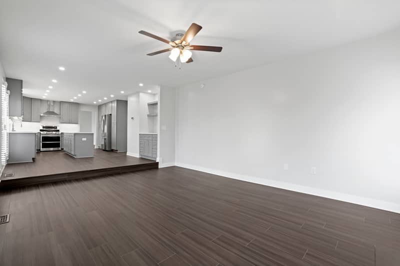 Family room with dark LVP flooring and ceiling fan looking toward the gray kitchen with stainless steel range, refrigerator, island, and built-in cabinetry