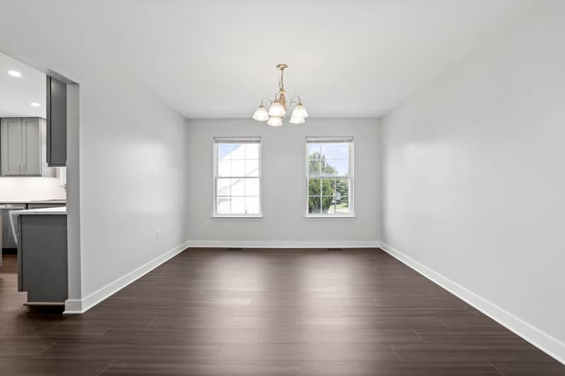 Empty dining room with dark LVP flooring, a chandelier, two windows overlooking the side yard, and an opening to the kitchen at left