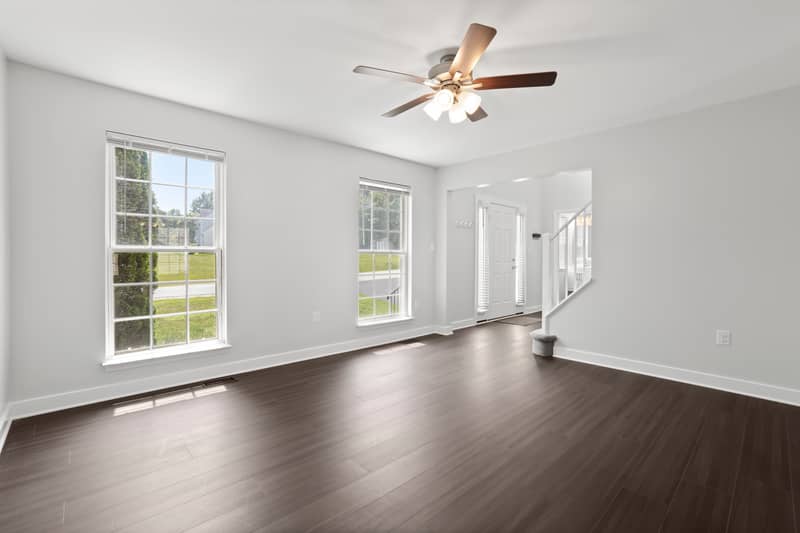 Empty living room with dark LVP flooring, ceiling fan, two large windows facing the front yard, and the front door with sidelight beside the carpeted staircase