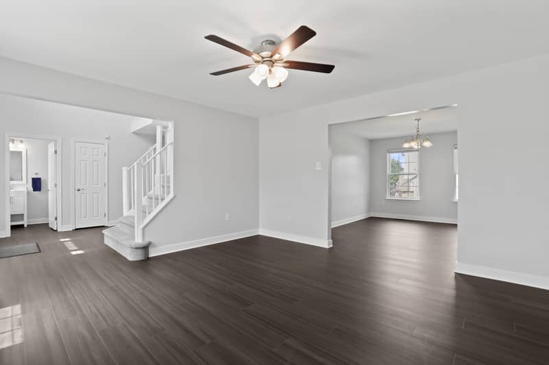 Living room with dark LVP flooring and ceiling fan, showing the carpeted staircase, a half bath and closet at left, and an opening to the chandelier-lit dining room at right