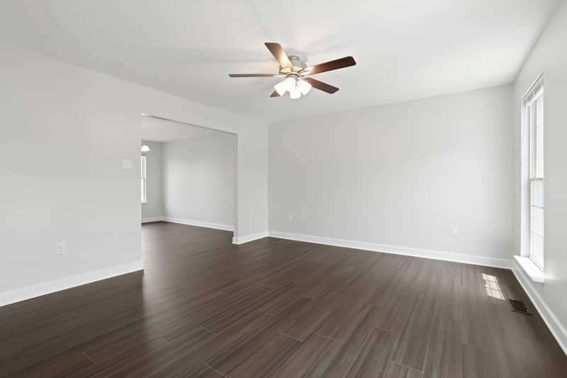 Empty living room with dark wood-look plank flooring, light gray walls, a ceiling fan, large window, and an open doorway to an adjoining room