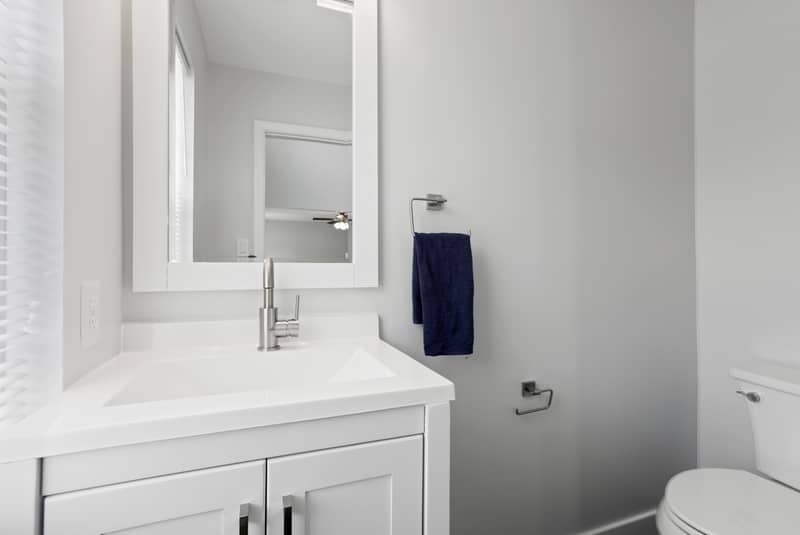 Small powder room with a white single-sink vanity, white quartz top, framed mirror, navy hand towel, and a toilet against light gray walls