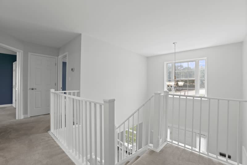 Carpeted second-floor landing with a white open stair railing, large window with chandelier above the stairwell, and doorways to bedrooms