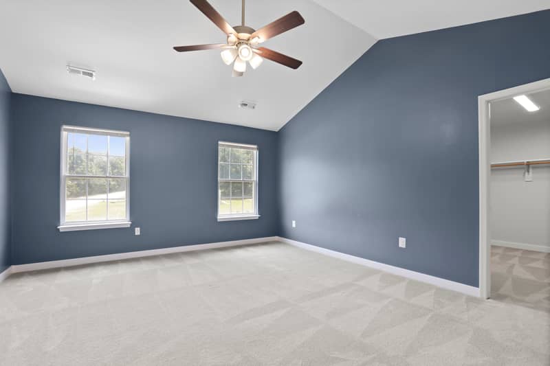 Spacious carpeted primary bedroom with blue-gray walls, vaulted ceiling, ceiling fan, two windows, and an open doorway to a walk-in closet