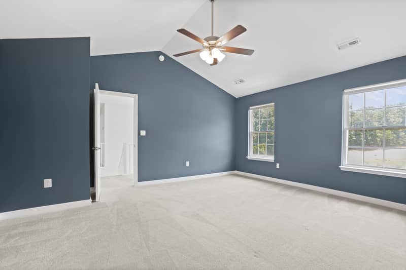 Empty carpeted primary bedroom with blue-gray walls, vaulted ceiling, ceiling fan, two windows overlooking trees, and an open door to the hallway