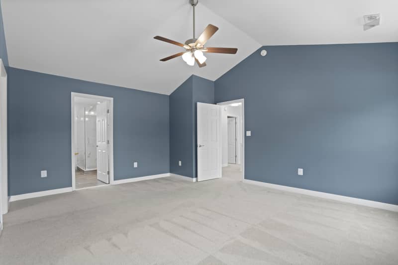 Carpeted primary bedroom with vaulted ceiling and ceiling fan, doorways revealing the tiled en-suite shower and the hallway beyond