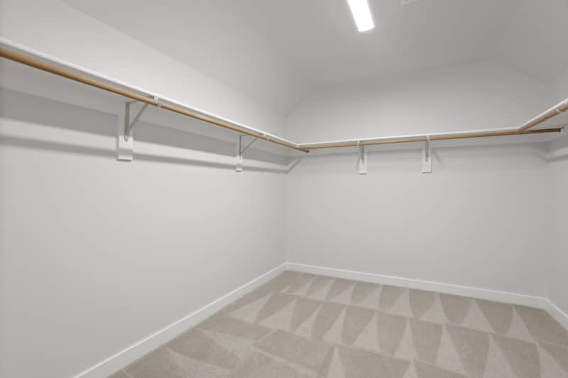 Empty carpeted walk-in closet with white walls, vaulted ceiling, overhead light, and wraparound shelves with hanging rods