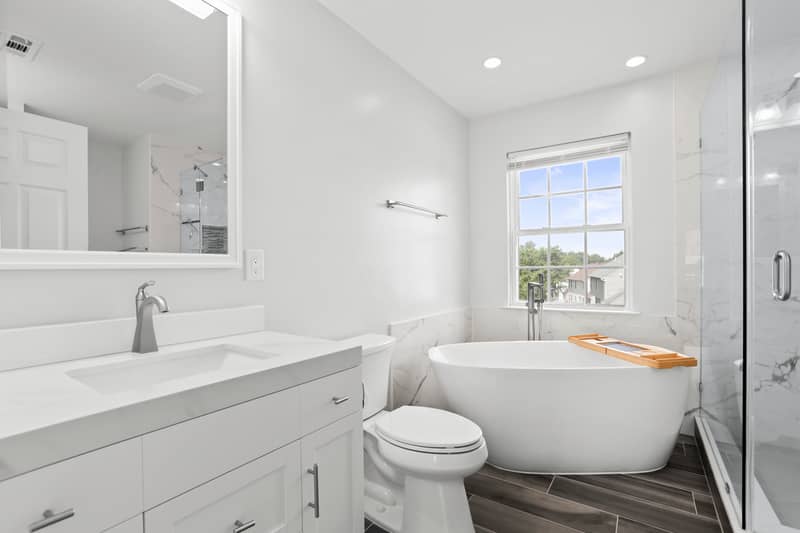Renovated primary bathroom with a freestanding soaking tub by a window, white quartz vanity, marble-look tile, glass walk-in shower, and wood-look tile floor