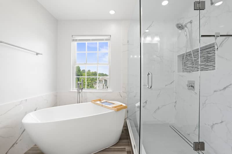 Primary bathroom with a freestanding white slipper tub beneath a window and a glass walk-in shower clad in marble-look tile with a tiled accent niche