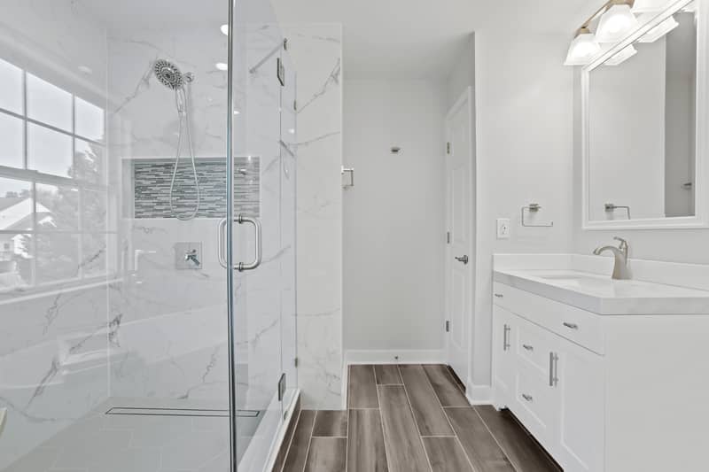 Primary bathroom showing a glass walk-in shower with marble-look tile, wood-look tile floor, and a white single vanity with quartz counter and large mirror