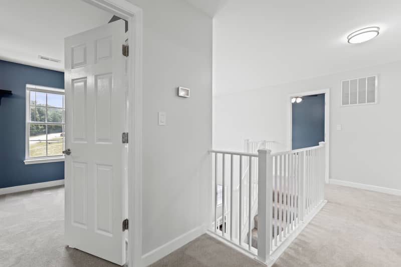 Carpeted upper-floor landing with a white-spindle stair railing overlooking the staircase and an open door into a blue-walled bedroom