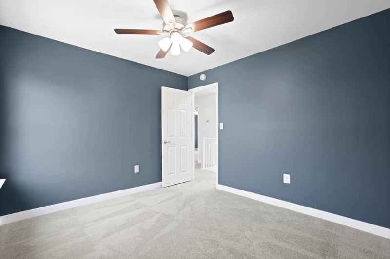 Slate-blue carpeted bedroom with a lighted ceiling fan and an open door leading to the upstairs landing